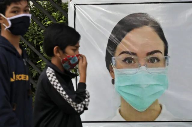 Indonesian youths walk past a banner that calls for people to always wear face mask as a precaution against the new coronavirus outbreak, in Jakarta, Indonesia, Wednesday, May 20, 2020. (Photo by Dita Alangkara/AP Photo)