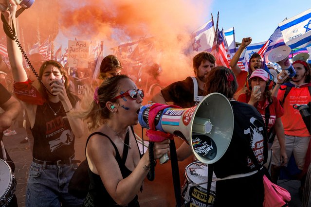 A demonstrator speaks on a megaphone as she marches through the smoke of a flare past others with Israeli and US flags during a protest against the Israeli government's judicial overhaul bill outside the US Embassy Tel Aviv branch office in Tel Aviv on July 11, 2023. Protesters blocked roads across Israel on July 11 hours after parliament adopted in a first reading a key clause of the government's judicial overhaul package which opponents say threatens democracy. (Photo by Menahem Kahana/AFP Photo)