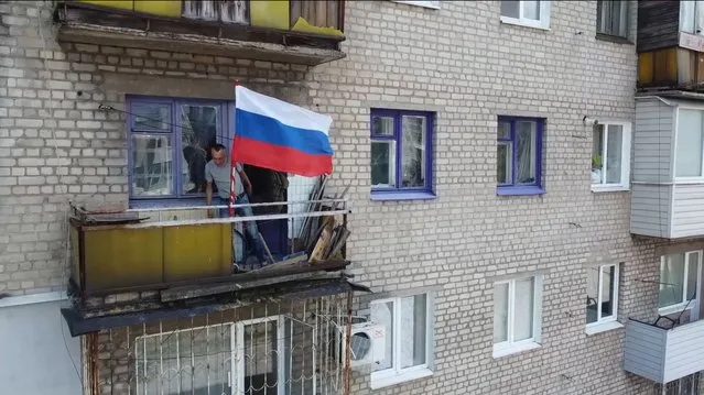 In this handout photo taken from video released by Russian Defense Ministry Press Service on Monday, July 4, 2022, a man sets a Russian national flag on a balcony of a residential building in Lysychansk, which is now territory under the Government of the Luhansk People's Republic control, eastern Ukraine. (Photo by Russian Defense Ministry Press Service via AP Photo)