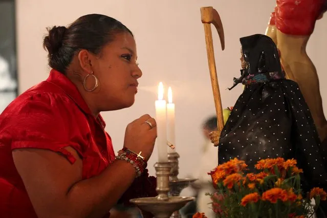 A woman blows smoke from a cigarette towards a figurine of La Santa Muerte (Saint Death) at a shrine during Day of the Dead celebrations in Ciudad Juarez, Mexico, November 2, 2015. The saint is often depicted as a skeletal "grim reaper" and followers leave offerings of tequila, rum, beer, cigarettes, cash, flowers and candy at altars adorned with rosaries and candles. (Photo by Jose Luis Gonzalez/Reuters)