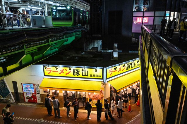 This photo taken on June 20, 2024 shows people queueing outside a ramen noodle shop below the tracks as a Yamanote Line train (top L) pulls into Gotanda station, one of 30 stations along the JR Yamanote Line in Tokyo. In service since 1885, the Yamanote Line is Tokyo's oldest, most important and most famous, with millions cramming onto the 35-kilometre (22-mile) route's distinctive green cars every day. (Photo by Richard A. Brooks/AFP Photo)