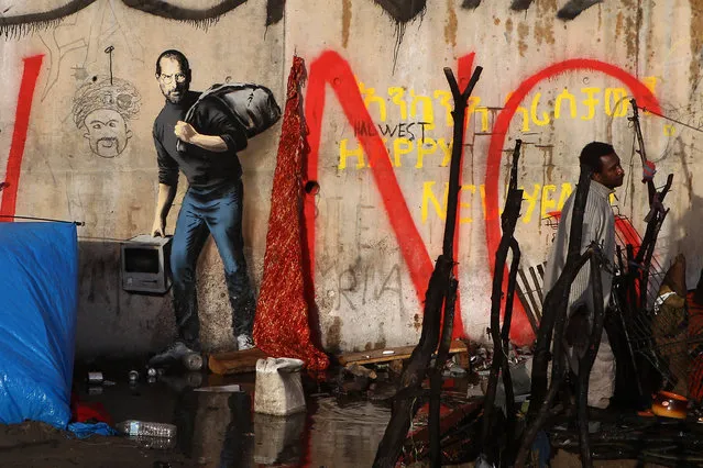 A painting by English graffiti artist Banksy is seen at the entrance of the Calais refugee camp in France, in Calais, northern France, Monday, December 21, 2015. The elusive graffiti artist has depicted the late Apple guru Steve Jobs – whose biological father was from Syria – carrying a black garbage bag and an early model of the Macintosh computer. (Photo by Michel Spingler/AP Photo)