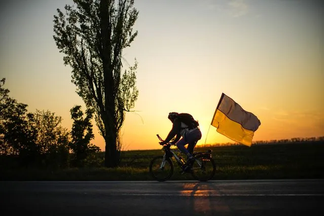 A man rides a bicycle with a Ukrainian flag on the road between Odesa and Mykolaiv, southern Ukraine, Saturday, May 14, 2022. (Photo by Francisco Seco/AP Photo)
