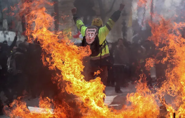 A Yellow Vest protester wearing a mask depicting the French President on which is written “psycho”, gestures behind flames rising from barricades, in Paris on March 16, 2019, during the 18th consecutive Saturday of demonstrations called by the “Yellow Vest” (gilets jaunes) movement. Demonstrators hit French city streets again on March 16, for a 18th consecutive week of nationwide protest against the French President's policies and his top-down style of governing, high cost of living, government tax reforms and for more “social and economic justice”. (Photo by Zakaria Abdelkafi/AFP Photo)