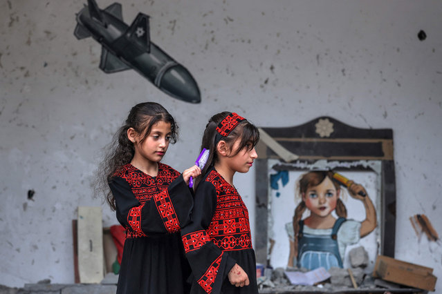 A Palestinian girl combs the hair of another as they pose before a mural during a mixed art exhibition titled “occupation kills childhood” taking place in the rubble of houses destroyed by Israeli air strikes during the May 2023 conflict in Deir al-Balah in the central Gaza Strip on June 8, 2023. (Photo by Mohammed Abed/AFP Photo)