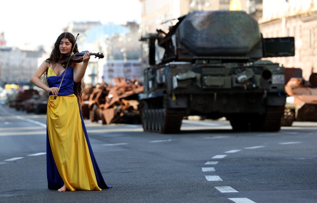 A woman dressed in the colors of the Ukrainian national flag takes part in a music video as she walks past damaged Russian armored military vehicles that were seized by the Ukrainian army amid the Russian invasion, along Khreshchatyk Street in Kyiv (Kiev), Ukraine on August 23, 2023. The damaged Russian military machinery was set up in downtown Kyiv prior to the upcoming Independence Day celebrations on 24 August. (Photo by Cathal Mcnaughton/EPA)