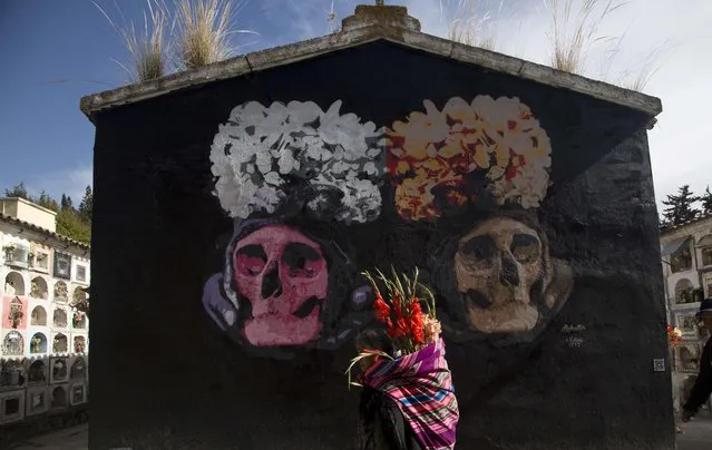 A woman walks past a mural depicting two human skulls or Natitas, taking flowers to a relative's grave in preparation for the upcoming Day of the Dead festivities, at the General Cemetery in La Paz, Bolivia, Tuesday, October 31, 2017. Day of the Dead or “Dia de los Muertos” is celebrated Nov. 1 and 2 to remember departed loved ones, many holding graveside picnics through the night. (Photo by Juan Karita/AP Photo)