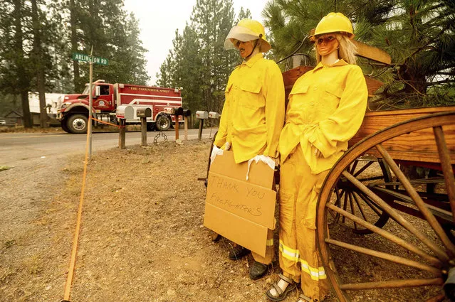 A fire truck battling the Dixie Fire passes mannequins dressed in firefighting gear in the Taylorsville community of Plumas County, Calif., on Saturday, August 14, 2021. The figures' owner said that he normally dresses the two up in tropical outfits and a fire crew dropped off firefighting apparel for them. (Photo by Noah Berger/AP Photo)