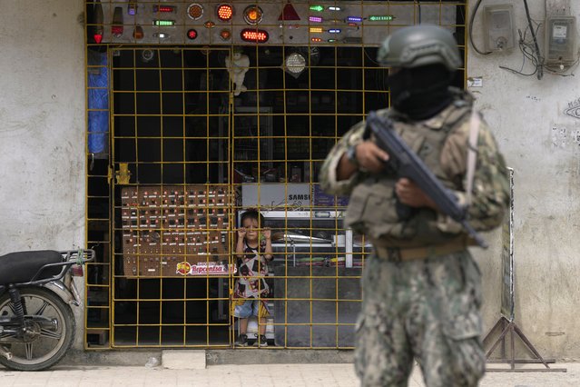 A boy watches from behind a storefront gate as a soldier stands guard at a security check point, in Duran, Ecuador, Monday, August 14, 2023. Ecuador's president declared a state of emergency in some areas after a presidential candidate was killed at a rally ahead of snap elections, set for Aug. 20. (Photo by Martin Mejia/AP Photo)