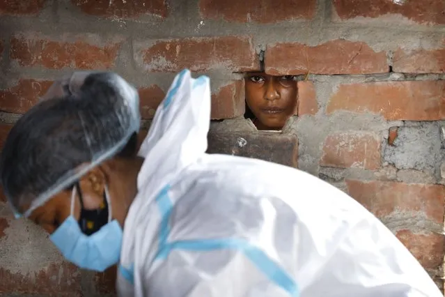 A girl looks out through a hole in the wall of her house as a health worker prepares for COVID-19 testing in Jamsoti village, Uttar Pradesh state, India, on June 9, 2021. India's vaccination efforts are being undermined by widespread hesitancy and fear of the jabs, fueled by misinformation and mistrust. That's especially true in rural India, where two-thirds of the country’s nearly 1.4 billion people live. (Photo by Rajesh Kumar Singh/AP Photo)
