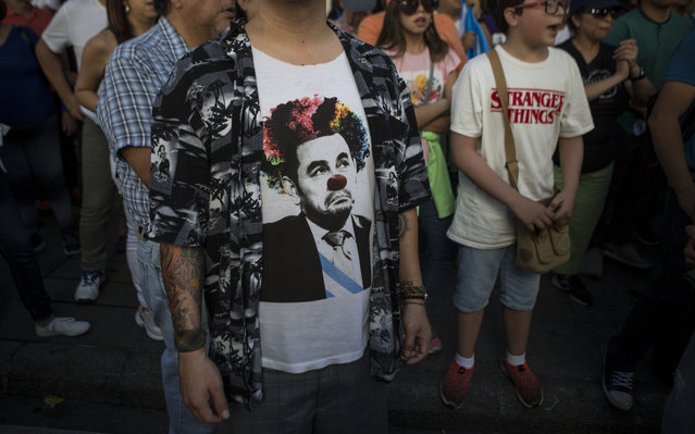 Demonstrators demand the resignation of Guatemalan President Jimmy Morales, one wearing a T-shirt of Morales as a clown, outside the Presidential House in Guatemala City, Saturday, July 27, 2019. Demonstrators are protesting an agreement their government signed with Washington to require migrants passing through the Central American country to seek asylum there, rather than pushing on to the U.S. Before becoming president, Morales was a professional comedian. (Photo by Oliver de Ros/AP Photo)