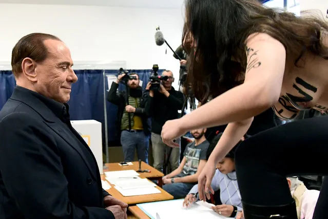 A woman alleged Femen activist jumps on a table in front of Silvio Berlusconi, leader of right-wing party Forza Italia, to protest topless with a bodypainting reading “Berlusconi, you have expired” on March 4, 2018 at a polling station in Milan. Italians vote today in one of the country's most uncertain elections, with far-right and populist parties expected to make major gains and Silvio Berlusconi set to play a leading role. (Photo by Miguel Medina/AFP Photo)