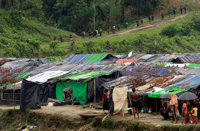 Rohingya refugees stand outside their temporary shelters at no man's land between Bangladesh-Myanmar border, as Myanmar security forces walk past a fence in Maungdaw, Myanmar September 9, 2017. (Photo by Danish Siddiqui/Reuters)