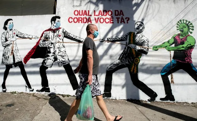 A man, wearing a protective face mask as a measure to curb the spread of the new coronavirus, walks past a mural depicting a tug-of-war between health workers and Brazil's President Jair Bolsonaro aided by a cartoon-styled coronavirus character, with a message that reads in Portuguese: “Which side are you on?”, in Sao Paulo, Brazil, Friday, June 19, 2020. (Photo by Andre Penner/AP Photo)