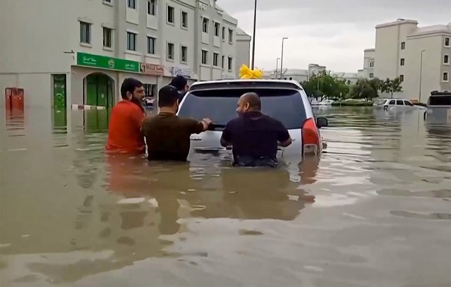 In this video grab from AFPTV, people push a stranded car along a flooded street in Dubai on April 16, 2024. Dubai, the Middle East's financial centre, has been paralysed by the torrential rain that caused floods across the UAE and Bahrain and left 18 dead in Oman on April 14 and 15. (Photo by Atif Bhatti/ESN via AFP Photo)
