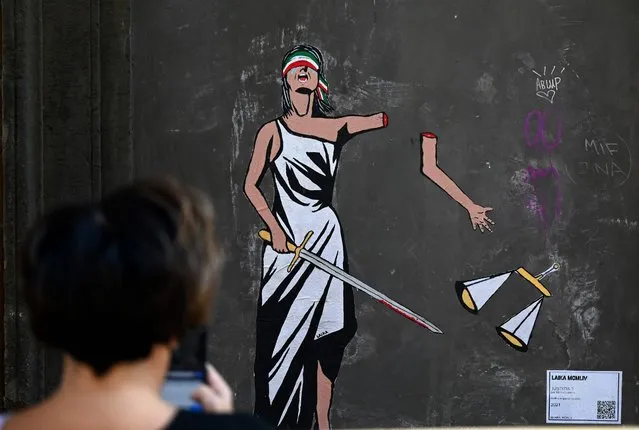 A woman takes a picture of a street art work by Italian street artist Laika, entitled “Iustitia” depicting the Greek goddess Dike, a divinity who represents justice, voluntarily amputating her left arm holding the famous scales, in downtown Rome on October 1, 2021 after pro-refugee Italian mayor Domenico Lucano was sentenced to 13 years for abetting illegal migration. (Photo by Filippo Monteforte/AFP Photo)