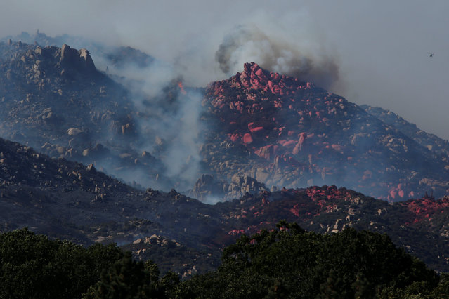 A smoke plume rises behind a rocky hillside covered with fire retardant as a firefighting helicopter flies during the Pilot Fire in San Bernardino county near the Deer Lodge Park area in Lake Arrowhead, California, U.S. August 9, 2016. (Photo by Patrick T. Fallon/Reuters)