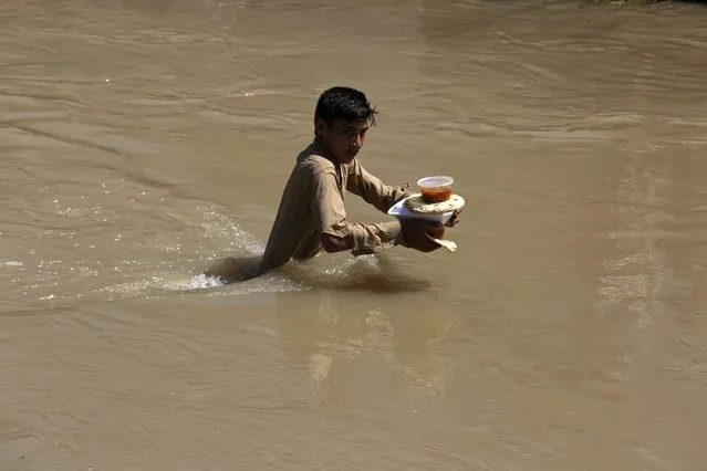 A displaced boy who fled his flood-hit home carries food rations as he wades through a flooded area on the outskirts of Peshawar, Pakistan, Sunday, August 28, 2022. Officials in Pakistan say deaths from widespread flooding have topped 1,000 since mid-June. Flash flooding from the heavy rains has washed away villages and crops as soldiers and rescue workers evacuated stranded residents to the safety of relief camps and provided food to thousands of displaced Pakistanis. (Photo by Mohammad Sajjad/AP Photo)