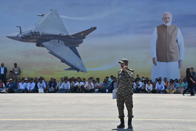 Visitors watch an aerial display seated under the shade of a poster during the 14th edition of Aero India 2023 at the Yelahanka Air Force Station in Bengaluru on February 14, 2023. (Photo by Manjunath Kiran/AFP Photo)