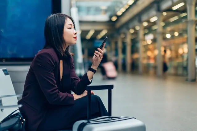 Beautiful Asian businesswoman working from mobile phone while waiting for boarding in airport / train station with luggage. (Phoot by Oscar Wong/Getty Images)