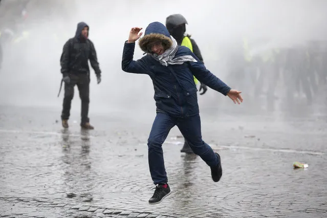 A demonstrator runs away after being sprayed with a water canon in clouds of teargas as yellow vest protesters clash with riot police on the famed Champs Elysees avenue in Paris, France, Saturday, January 12, 2019. Authorities deployed 80,000 security forces nationwide for a ninth straight weekend of anti-government protests. (Photo by Thibault Camus/AP Photo)