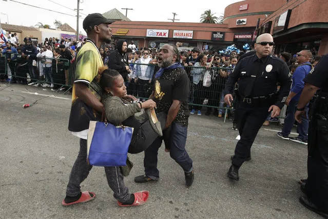 Two men carry a woman injured in a stampede as people gather to watch a hearse carrying the casket of slain rapper Nipsey Hussle Thursday, April 11, 2019, in Los Angeles. The 25-mile procession traveled through the streets of South Los Angeles after a memorial service, including a trip past Hussle's clothing store, The Marathon, rear, where he was gunned down March 31. (Photo by Jae C. Hong/AP Photo)