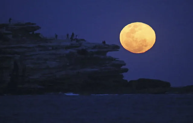 The moon rises with an orange glow as people watch from a rock cliff at Bondi Beach in Sydney, Tuesday, September 9, 2014. The supermoon, known to scientists as a perigee moon, occurs when Earth's neighbor appears at its largest and brightest compared to other full moons when it orbits closer to our planet. (Photo by Rick Rycroft/AP Photo)