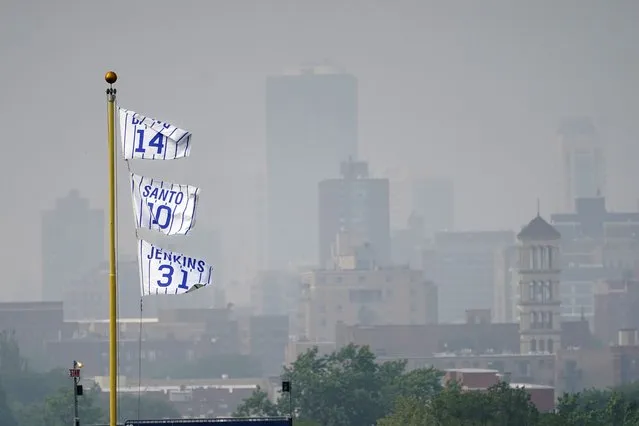 The flags of Chicago Cubs' Hall of Fame players Ernie Banks (14) Ron Santo (10) and Ferguson Jenkins fly off the Wrigley Field left field foul pole as a veil of haze from Canadian wildfires shroud high rise buildings along Lake Michigan before a baseball game between the Cubs and the Philadelphia Phillies Tuesday, June 27, 2023, in Chicago. (Photo by Charles Rex Arbogast/AP Photo)