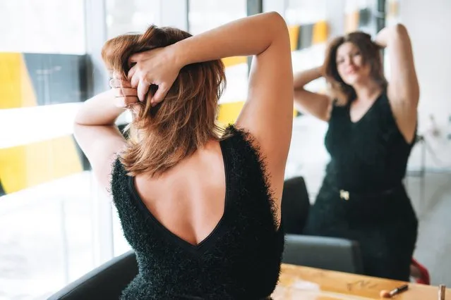 Beautiful smiling young woman plus size body positive in black evening dress make hair style near mirror at beauty studio. (Photo by Galina Zhigalova/Getty Images)