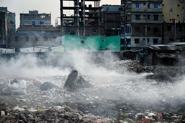 A woman scouts for reusable items from a pile of trash as smoke rises from a nearby area  in Dhaka on January 31, 2022. (Photo by Munir Uz Zaman/AFP Photo)