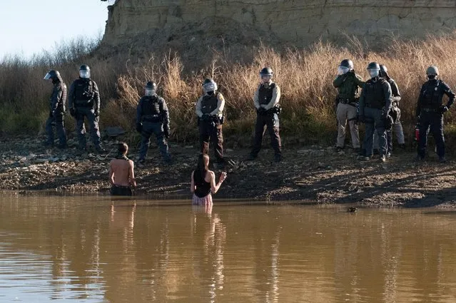Two people stand in the water of a river while police officers guard the shore during a protest against the building of a pipeline near the Standing Rock Indian Reservation near Cannonball, North Dakota, U.S. November 2, 2016. (Photo by Stephanie Keith/Reuters)