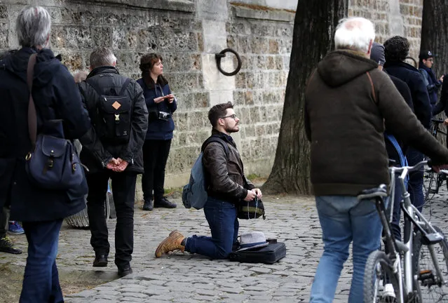 Man kneels as people came to watch and photograph the Notre Dame cathedral after the fire in Paris, Tuesday, April 16, 2019. Experts are assessing the blackened shell of Paris' iconic Notre Dame cathedral to establish next steps to save what remains after a devastating fire destroyed much of the almost 900-year-old building. With the fire that broke out Monday evening and quickly consumed the cathedral now under control, attention is turning to ensuring the structural integrity of the remaining building. (Photo by Christophe Ena/AP Photo)