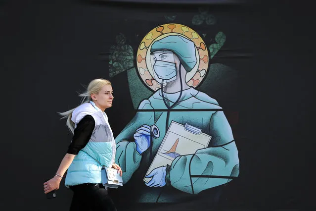A woman walks by a depiction of a medical staff wearing protective equipment, executed in the style of orthodox icons, in Bucharest, Romania, Wednesday, April 29, 2020. The artwork, among others depicting medical staff in the manner of religious icons, created by designer Wanda Hutira, is part of a campaign called Thank You Doctors, meant to raise awareness to the work of medical staff fighting the COVID-19 pandemic. Following public pressure by Romania's influential Orthodox church the artworks, described as “blasphemous” will be removed from all locations in the Romanian capital, according to the agency behind the project. (Photo by Vadim Ghirda/AP Photo)