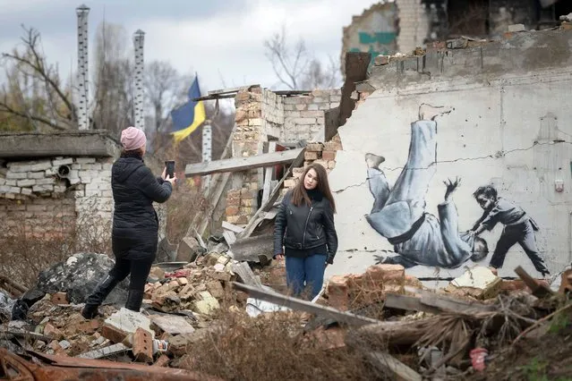 Women take a picture with an artwork on a building destroyed by fighting in Borodyanka, Kyiv region, Ukraine, Sunday, November 13, 2022. The painting appears to have been made by British street artist Banksy, who confirmed on his Instagram account that he made another mural in the village that depicts a gymnast. (Photo by Andrew Kravchenko/AP Photo)