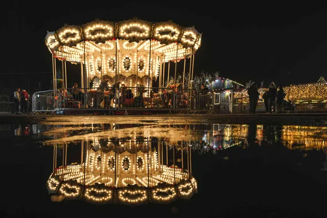 A carousel is reflected in a puddle at a Christmas fair in Bucharest, Romania, Saturday, November 27, 2021. The Romanian capital will have three Christmas fairs open for public in the coming weeks and access to the venues will be conditioned by a COVID-19 green pas, proving the holder's vaccination or recovery after the infection. (Photo by Vadim Ghirda/AP Photo)