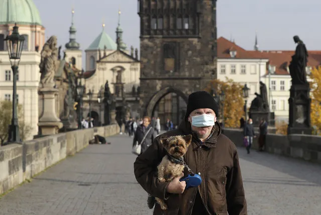 A man wearing a face mask carries his dog across the medieval Charles Bridge in Prague, Czech Republic, Wednesday, October 21, 2020. In another desperate attempt to slow the rise of coronavirus infections in the Czech Republic, Health Minister Roman Prymula has announced a ban on free movement of people in the country and a closure of many stores, shopping malls and hotels. At the same time, state offices will limit their opening hours. Prime Minister Andrej Babis says those measures should prevent the collapse of the health system in early November. (Photo by Petr David Josek/AP Photo)