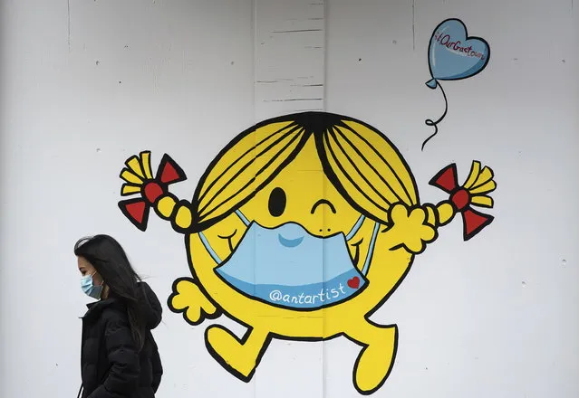 A woman wearing a face mask walks past artwork of author and illustrator Roger Hargreaves' 'Little Miss Sunshine' character wearing a face mask, painted on the outside of a boarded up business in downtown Vancouver, on Sunday, April 19, 2020. (Photo by Darryl Dyck/The Canadian Press via AP Photo)