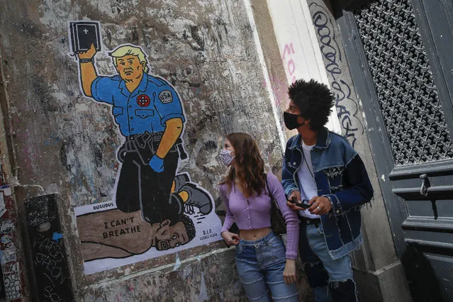 People pose next to an artwork by French artist Dugudus depicting U.S. President Donald Trump as a police officer pressing his knee into the neck of George Floyd while holding a bible, in Paris, France, Saturday, June 6, 2020. (Photo by Francois Mori/AP Photo)