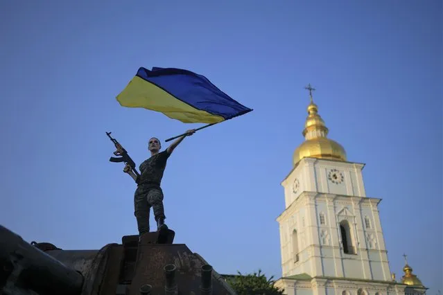 A woman brandishes the Ukrainian flag on top of a destroyed Russian tank in Kyiv, Ukraine, Friday, June 10, 2022. With war raging on fronts to the east and south, the summer of 2022 is proving bitter for the Ukrainian capital, Kyiv. The sun shines but sadness and grim determination reign. (Photo by Natacha Pisarenko/AP Photo)