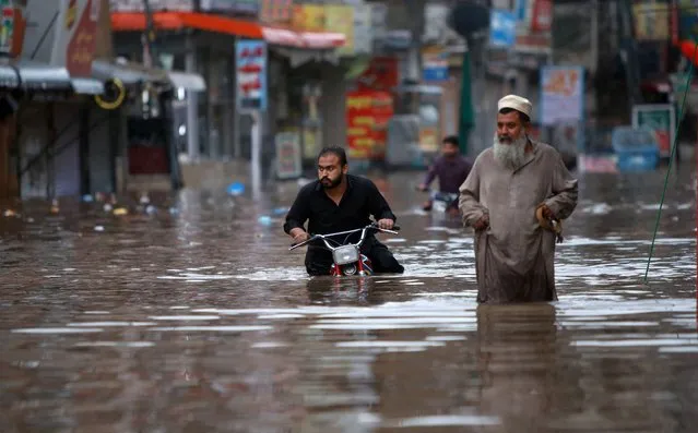 A man wades next to another pushing a motorbike along a waterlogged street after the monsoon rainfall in Rawalpindi on July 13, 2022. (Photo by Raja Imran/AFP Photo)