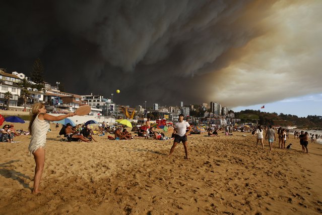 Vacationers play paddle ball on the beach backdropped by a darkening sky caused by smoke from nearby forest fires in Viña del Mar, Chile, Friday, February 2, 2024. (Photo by Martin Thomas, Aton Chile via AP Photo)