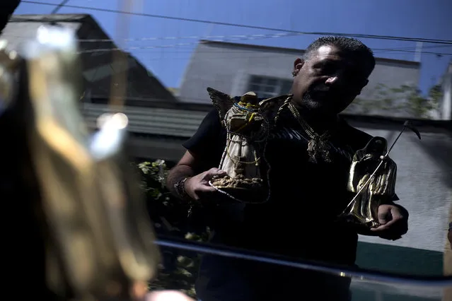 In this March 1, 2017 photo, Manuel Zavala, a devotee of Death Saint or “Santa Muerte”, is reflected on a car window as he holds statues of the folk saint during a pilgrimage in Mexico City's Tepito neighborhood. Zavala credits the saint for turning him around: “Thanks to a person I love a lot, my White Girl, my life has changed and now I'm not the second-rate guy I was before”. (Photo by Marco Ugarte/AP Photo)