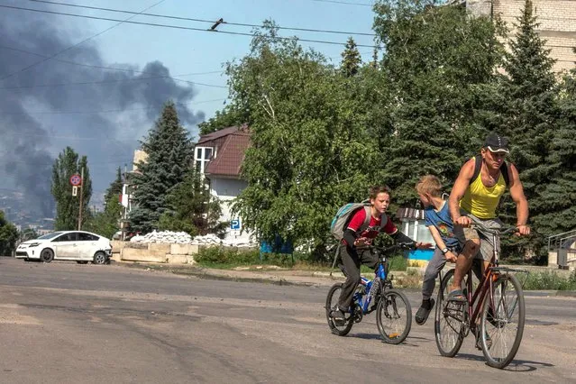 A local man with kids rides a bicycle along an empty street, as smoke rises after shelling in the background, amid Russia's attack on Ukraine, in the town of Lysychansk, Luhansk region, Ukraine on June 10, 2022. (Photo by Oleksandr Ratushniak/Reuters)