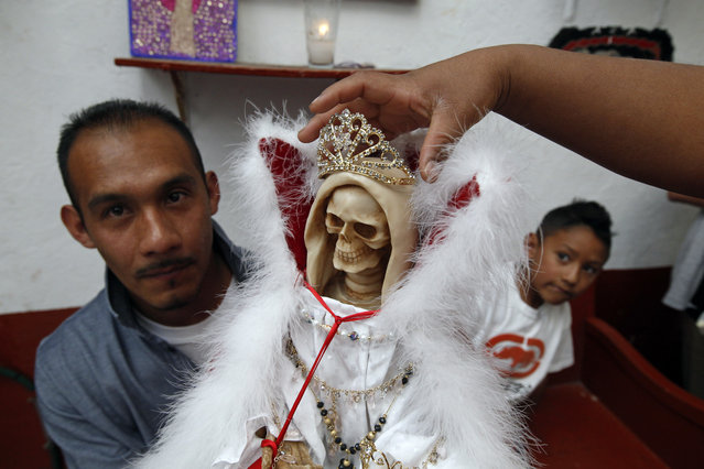 In this February 19, 2017 photo, a man holds a Death Saint statue as his wife places a crown and his son looks on, at Mercy Church on the edge of Mexico City's Tepito neighborhood. To followers, she's known as the Death Saint, the White Girl, the Skinny One, or just Sister – and a life-transforming answer to their prayers. To the Vatican, though, she's an irritation seen as leading the faithful astray. (Photo by Marco Ugarte/AP Photo)