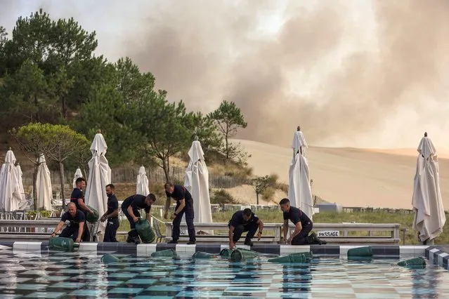 Firefighters put gas in safety in the swimming pool of the 5 star hotel La Corniche in the town of Pilat sur Merle on July 18, 2022, South West of France, as smoke of a fire in La Teste de Buch is seen in background. In scorching heat, with more than 40°C, some 8,000 people had to leave – in a “preventive manner” according to the prefecture – the Miquelots and Pyla-sur-Mer, districts of the municipality of La Teste-de-Buch, a town of 28,000 inhabitants where 4,300 hectares of forest went up in smoke. (Photo by Thibaud Moritz/AFP Photo)