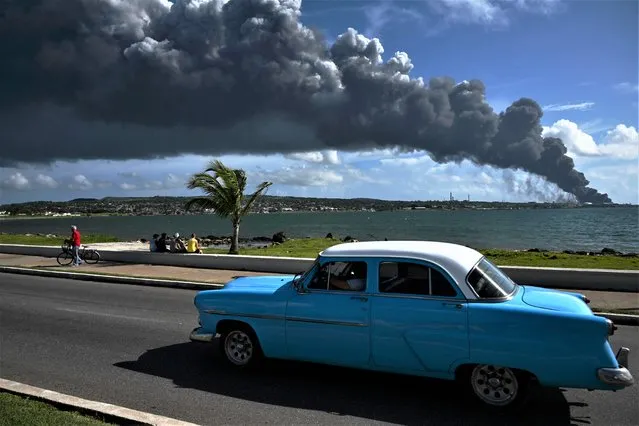 Black smoke from an oil tank on fire is seen while an old American car passes by a street in Matanzas, Cuba, on August 6, 2022. The fire caused by lightning on Friday in a fuel depot in Matanzas, in western Cuba, spread to a second tank at dawn this Saturday and caused 49 injuries, official sources reported. (Photo by Yamil Lage/AFP Photo)