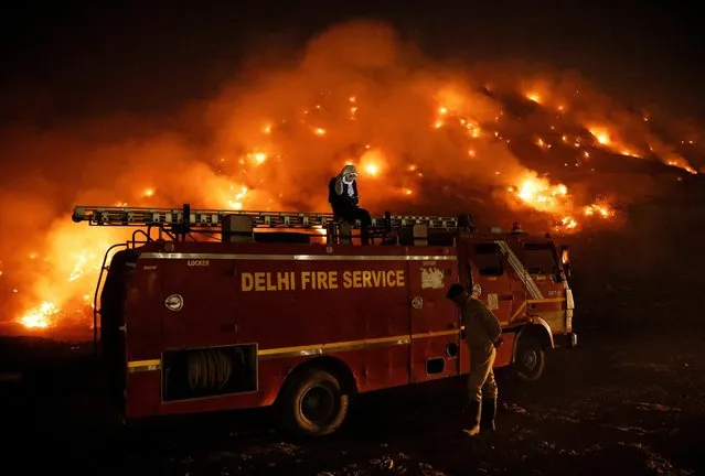 A firefighter uses his mobile phone as he sits on top of a fire truck as smoke billows from burning garbage at the Bhalswa landfill site in New Delhi, India, April 27, 2022. (Photo by Adnan Abidi/Reuters)