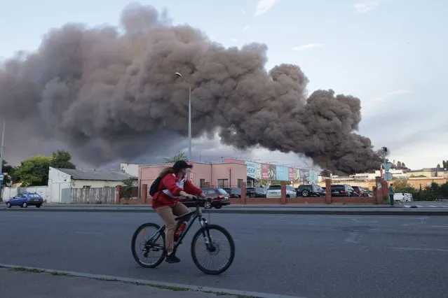 A woman riding a bicycle drives past a cloud of smoke from a fire in the background after a missile strike on a warehouse of an industrial and trading company in Odessa on July 16, 2022, amid the Russian invasion of Ukraine. (Photo by Oleksandr Gimanov/AFP Photo)