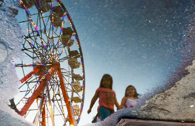 A colorful ferris wheel and two girls are reflected in a puddle on Tuesday, June 3, 2014 in Langley Park Maryland at a carnival sponsored by The Langley Park Boys and Girls Club. It is located near New Hampshire Ave. and Riggs Road and is running until June 15th. (Photo by Sarah L. Voisin/The Washington Post)