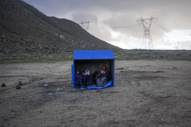 Asylum-seeking migrants from Peru and Ecuador take shelter from the wind and rain as they wait to be processed in a makeshift, mountainous campsite after crossing the border with Mexico, Friday, February 2, 2024, near Jacumba Hot Springs, Calif. (Photo by Gregory Bull/AP Photo)